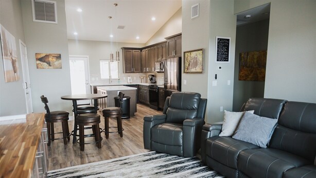 A modern kitchen with dark cabinets, a central island, and stainless steel appliances. There is a dining area with a wooden table and bar stools. A leather sofa and a patterned rug are in the living area. - Cougar Canyon Unit A (Heber-Overgaard)