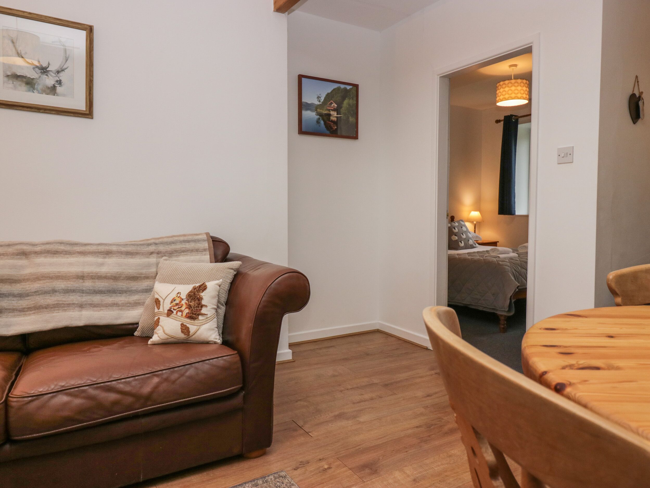 A living room with a brown leather sofa, a wooden dining table, and a bed in the adjacent room.