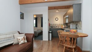 Un salon chaleureux avec un canapé en cuir marron, une table à manger en bois avec des chaises, un coin cuisine doté de meubles gris et d’un four à micro-ondes, et un couloir menant à une chambre.