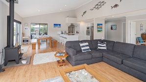 A spacious living room with a grey sofa, wooden coffee table, and a fireplace. There is a dining area with a table and chairs, and a kitchen in the background.