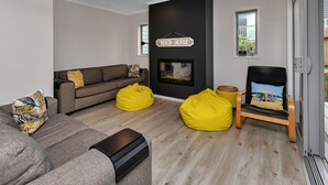 A modern living room with a fireplace, a sofa, two yellow bean bags, and a chair with a beach-themed cushion. - Seaside Escape - Ruakaka Holiday Home (Ruakaka)