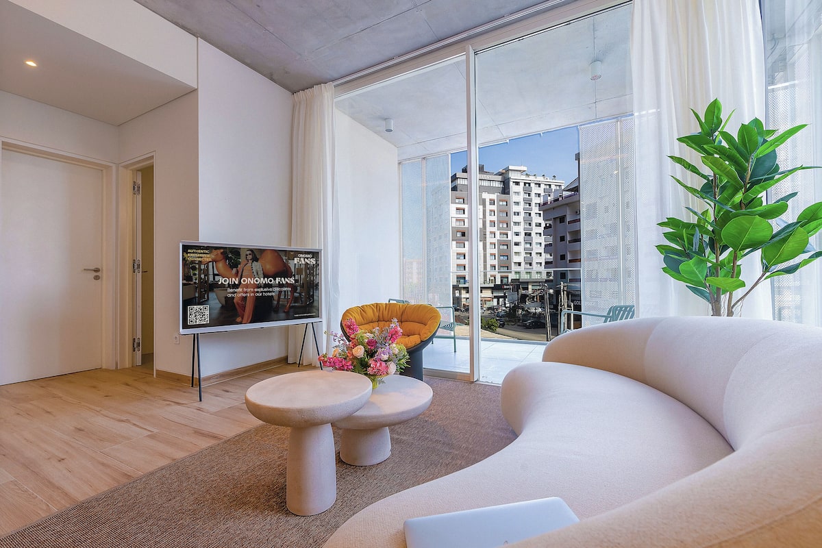 Un salon moderne avec un canapé, une table basse et un téléviseur. Une fenêtre offre une vue sur les bâtiments à l’extérieur.
