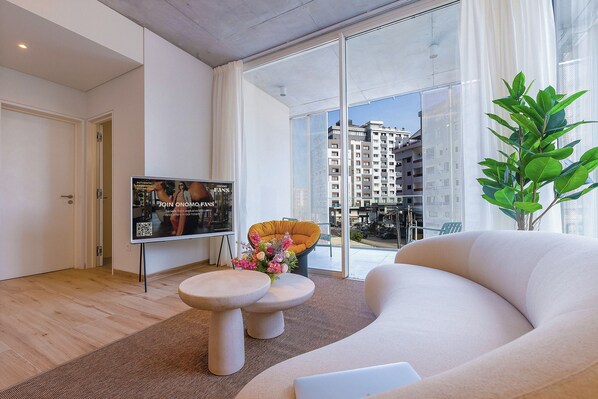 Una sala de estar moderna con un sofá, una mesa de centro y un televisor. Se ve una ventana con vistas a edificios.