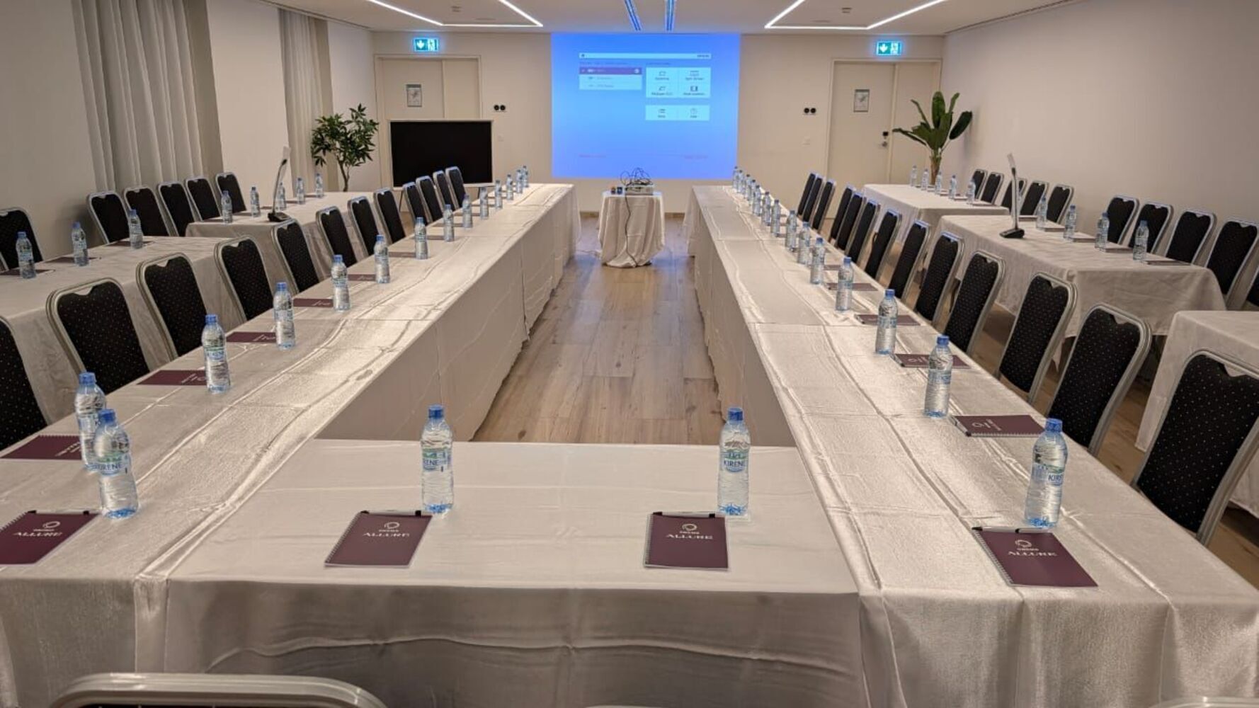A conference room with long tables, water bottles, and nameplates, equipped with a projector screen at the front.