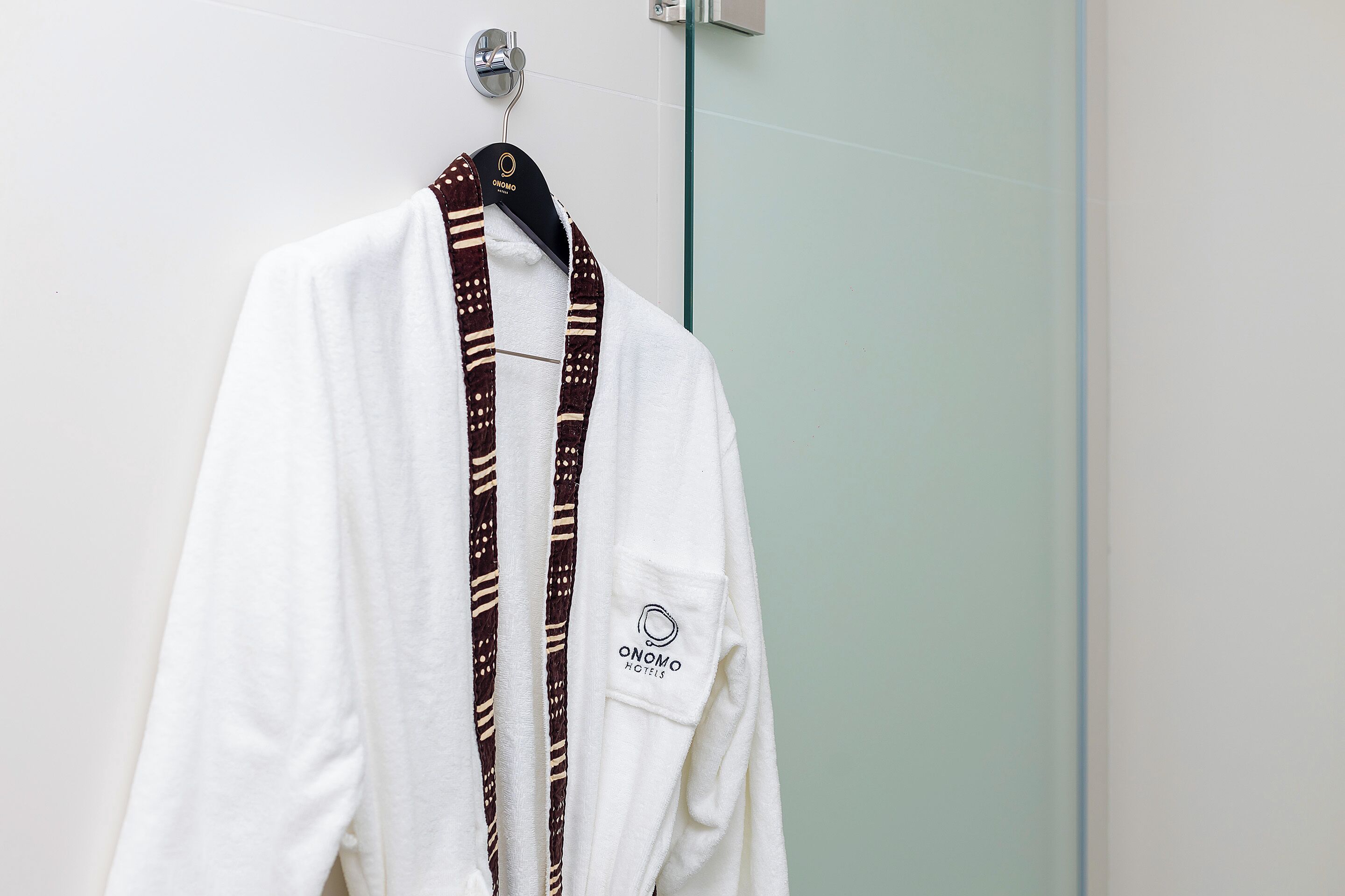 Una bata blanca con una bufanda marrón estampada colgada de un perchero en un baño con puerta de ducha de vidrio.
