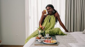 A person sitting on a bed with a tray of food and a drink, a vase with flowers, and a window with curtains.