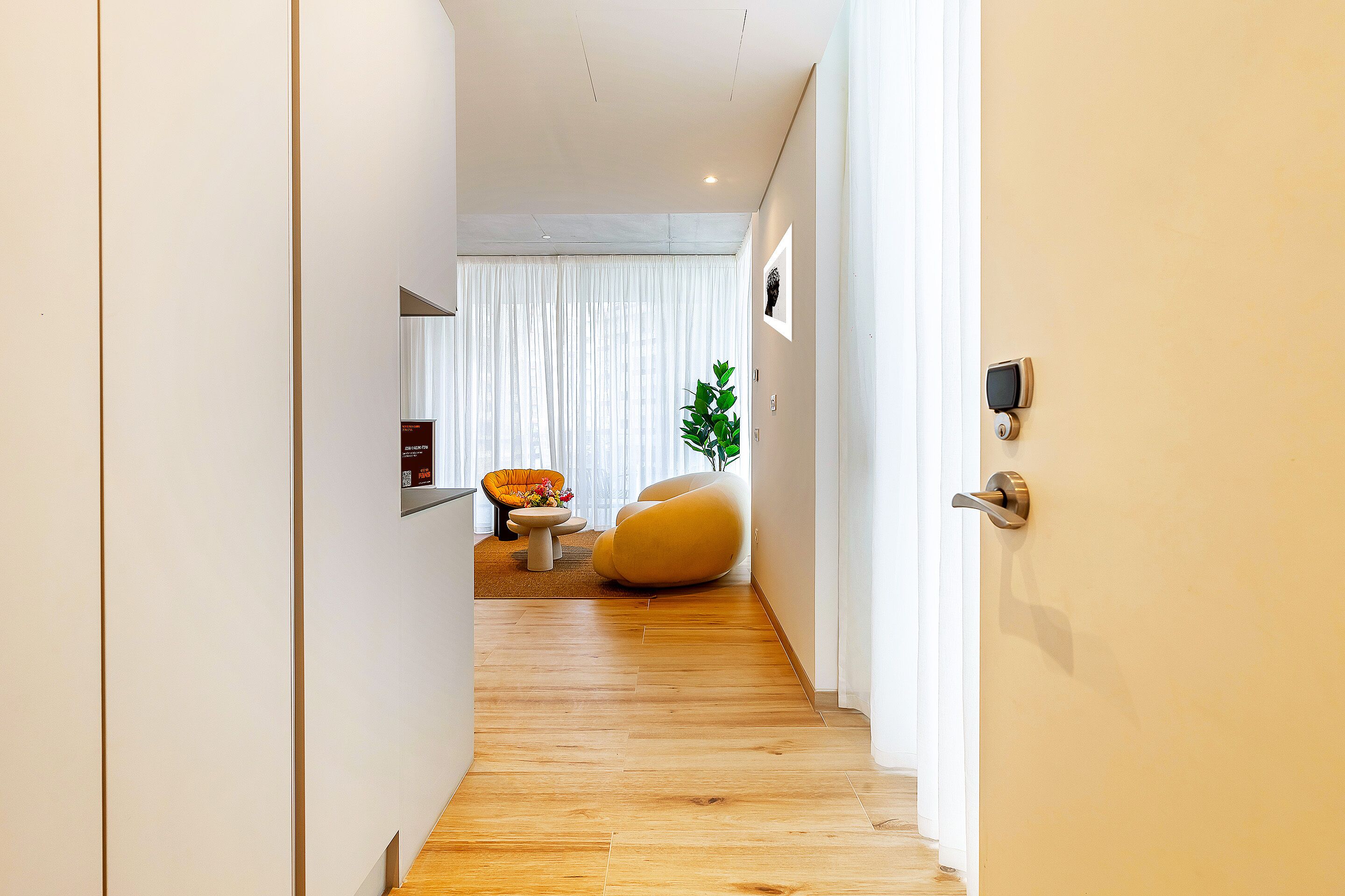 Un pasillo moderno con piso de madera, una zona de estar con una silla amarilla y una planta, y una puerta con un dispositivo de seguridad.