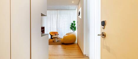 Un pasillo moderno con piso de madera, una zona de estar con una silla amarilla y una planta, y una puerta con un dispositivo de seguridad.