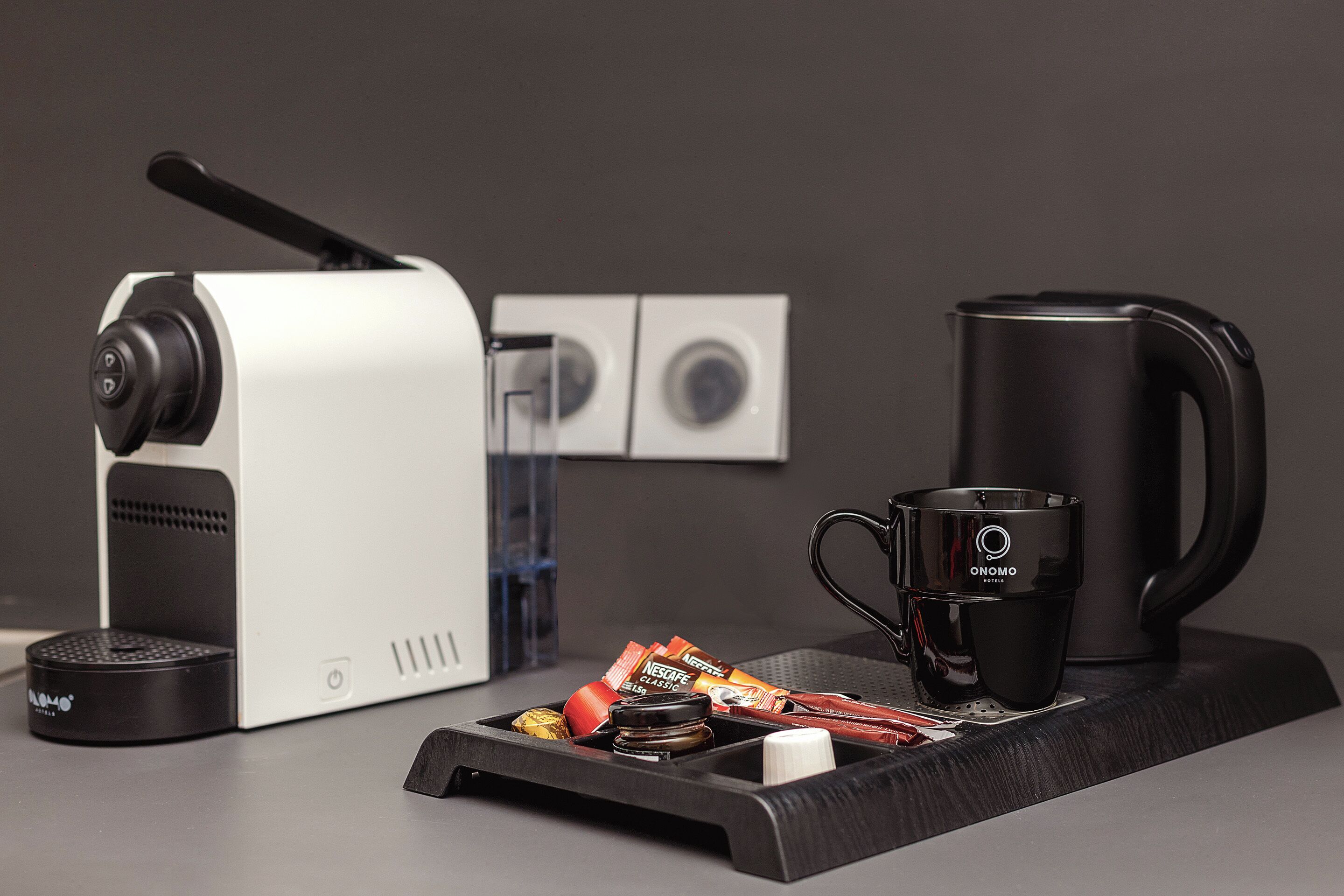 Eine Kaffeemaschine, eine schwarze Tasse, ein Wasserkocher und Kaffeepads auf einem Tablett.