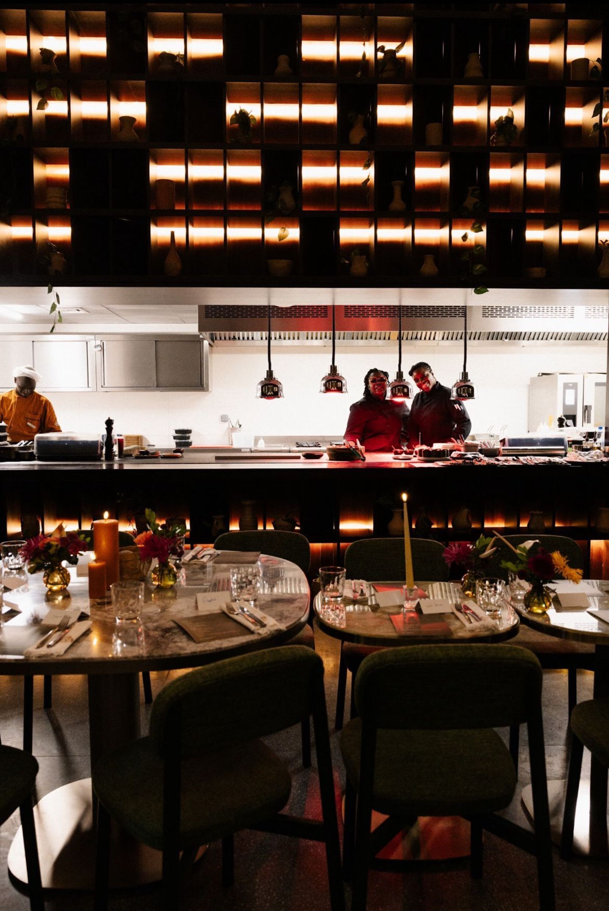 A restaurant with a long counter, multiple tables, and chairs. There are hanging lights above the counter, and a person is visible in the background.