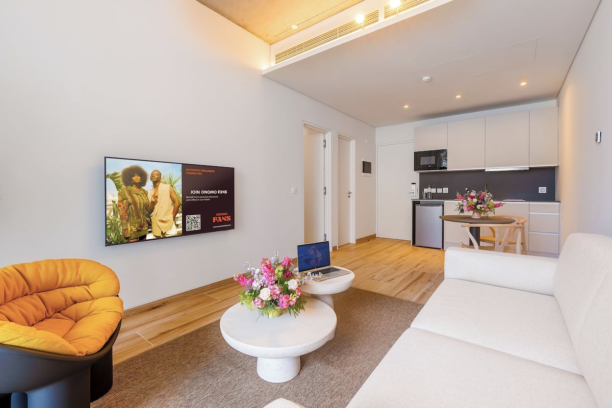 Un salon moderne avec un canapé, un fauteuil jaune, une table basse ornée de fleurs, un ordinateur portable posé sur une petite table, et une cuisine en arrière-plan.