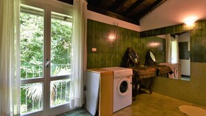 A room with a washing machine, a sink, and a mirror. There is a window with curtains and a view of greenery outside. - Beautiful private villa for 6 guests with WIFI, A/C, TV, patio and pets allowed (Cosio Valtellino)