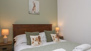 A neatly made bed with a green quilt, two pillows with animal prints, and a wall-mounted painting of a rabbit. - Winder Green (Penrith)