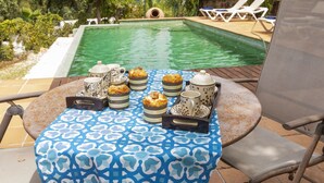 Una mesa junto a la piscina con mantel estampado, una bandeja de pasteles y una tetera.
