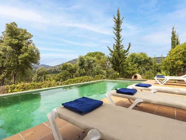 An infinity pool with lounge chairs and towels, surrounded by lush greenery and hills in the background.