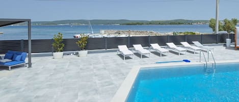 Piscina no terraço com espreguiçadeiras e área de estar, com vista para um corpo d'água e terra distante.
