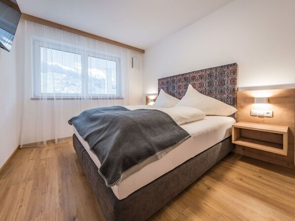 A modern bedroom with a large bed, a bedside table with a lamp, and a window with sheer curtains.