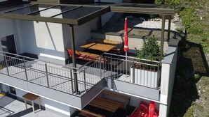 A modern outdoor patio with a glass enclosure, wooden furniture, and red chairs.