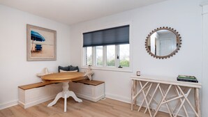 Ein Zimmer mit einem runden Holztisch, einer Bank, einem gerahmten Gemälde, einem Fenster mit Verdunkelungsvorhang, einem runden Spiegel mit Zierrahmen und einem kleinen weißen Konsolentisch mit Büchern.