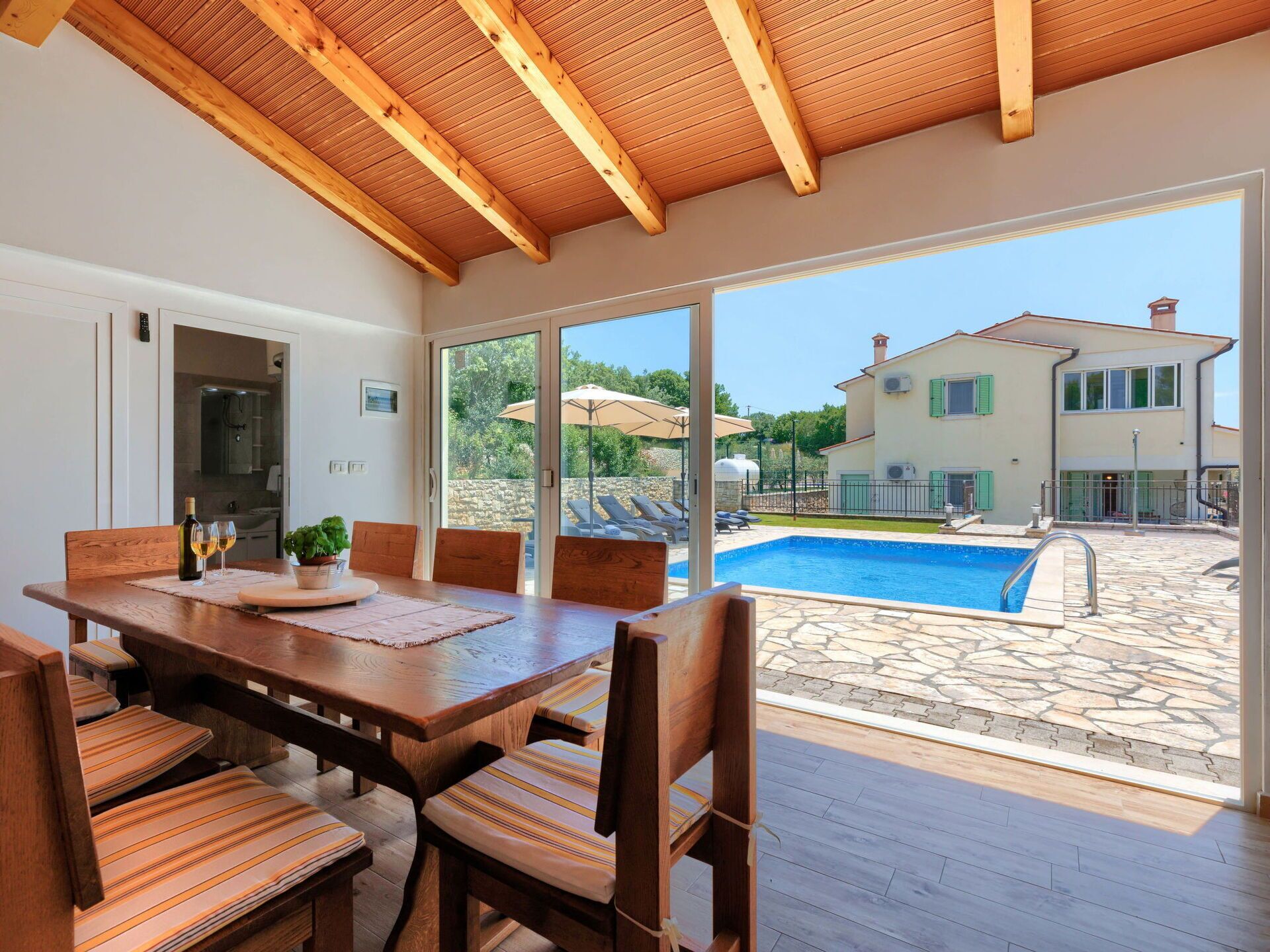 A spacious dining area with a wooden table and chairs, a view of a swimming pool, and a building with a stone facade in the background.