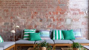 A cozy seating area with a brick wall, a sofa with striped cushions, and wooden tables with potted plants. - Wonderful  villa for 7 people with private pool, A/C, WIFI, TV and patio (Malgrat De Mar)