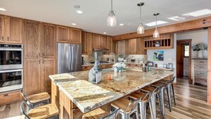 A modern kitchen with wooden cabinets, stainless steel appliances, a large granite island with bar stools, pendant lighting, and a view of the living area.