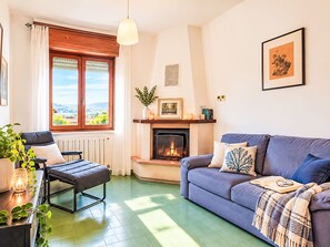 Un salon chaleureux avec une cheminée, un canapé, un fauteuil et une vue sur l’extérieur.