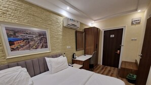 Una habitación de hotel con cama, escritorio, silla, aire acondicionado, un cuadro en la pared y una puerta.