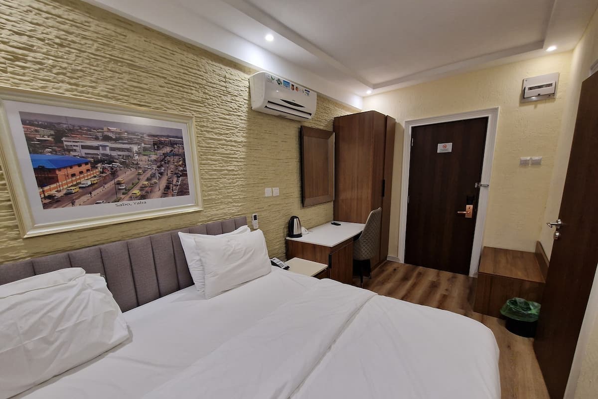A hotel room with a bed, a desk, a chair, an air conditioner, a framed picture on the wall, and a door.