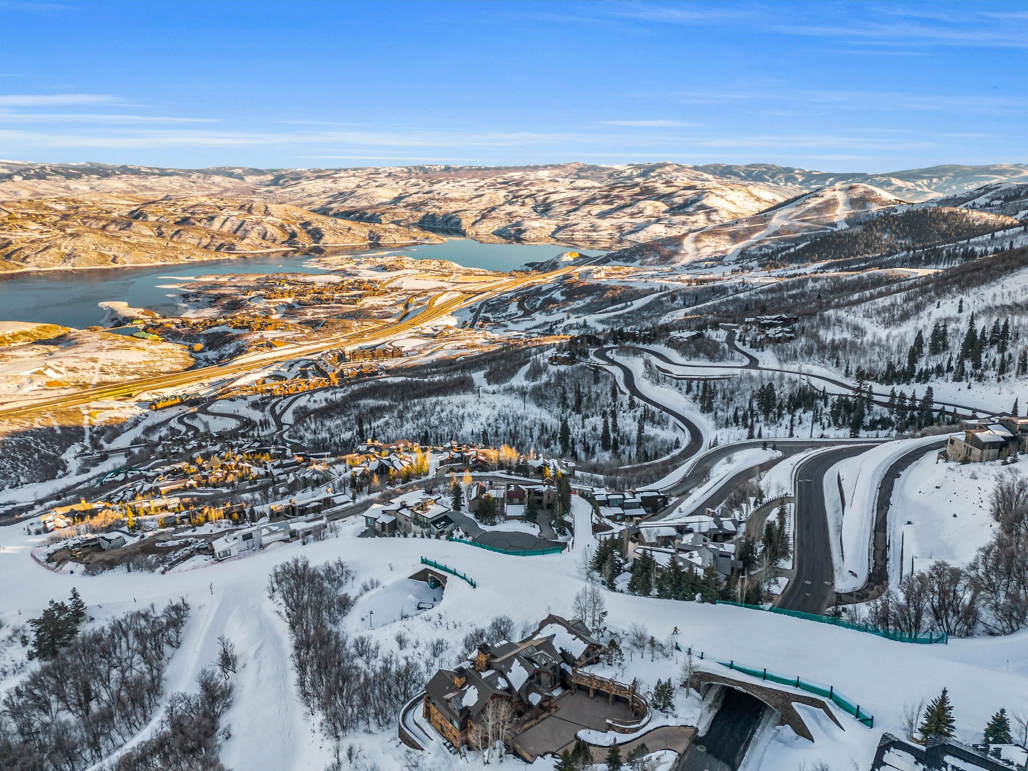 Paisagem coberta de neve com uma estrada sinuosa, um pequeno povoado com edifícios e uma grande massa de água ao fundo.