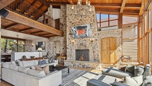Amplia sala de estar con chimenea de piedra, techo alto con vigas de madera y balcón con vistas al paisaje.