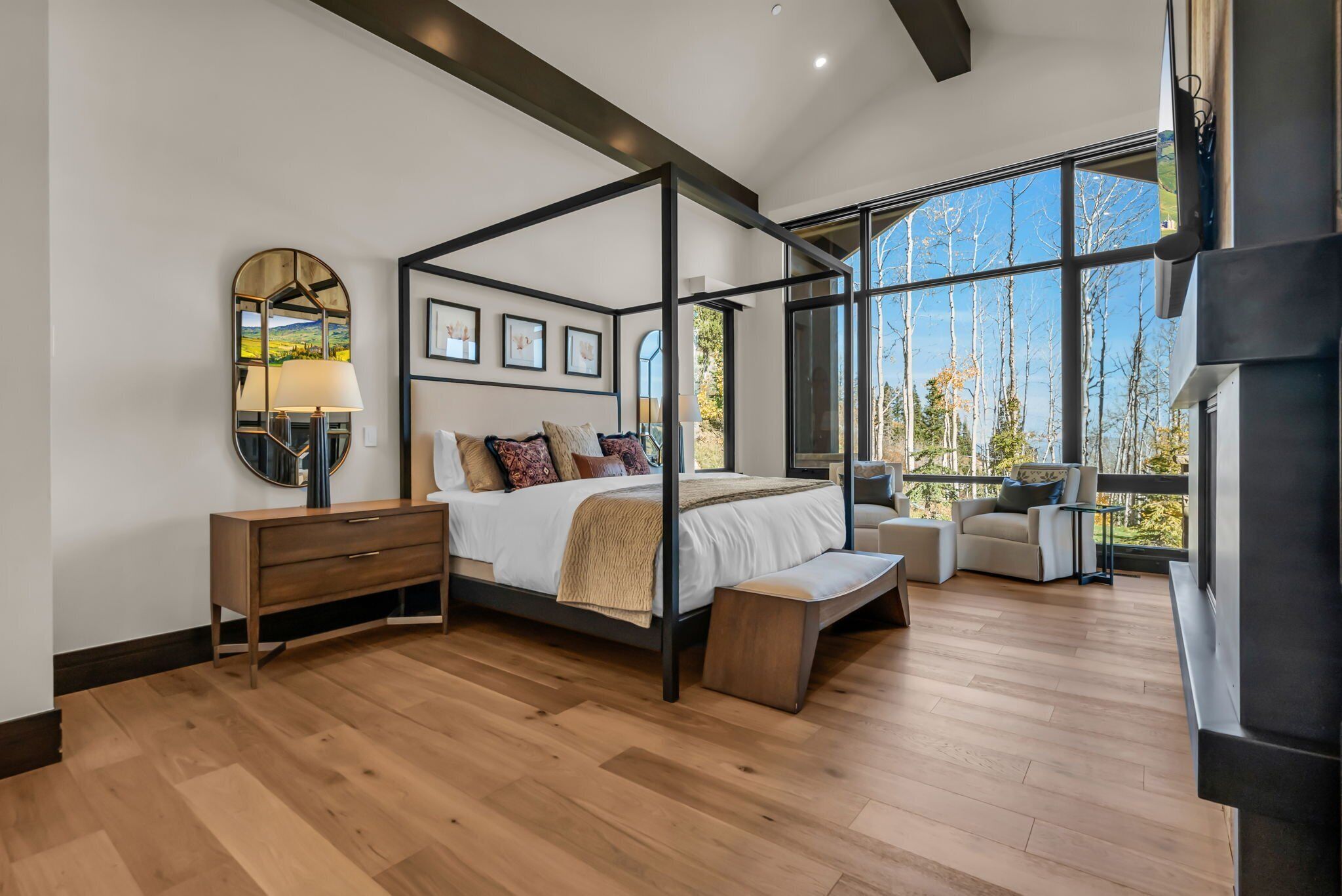 A modern bedroom with a four-poster bed, a large mirror, and a view of trees through large windows.