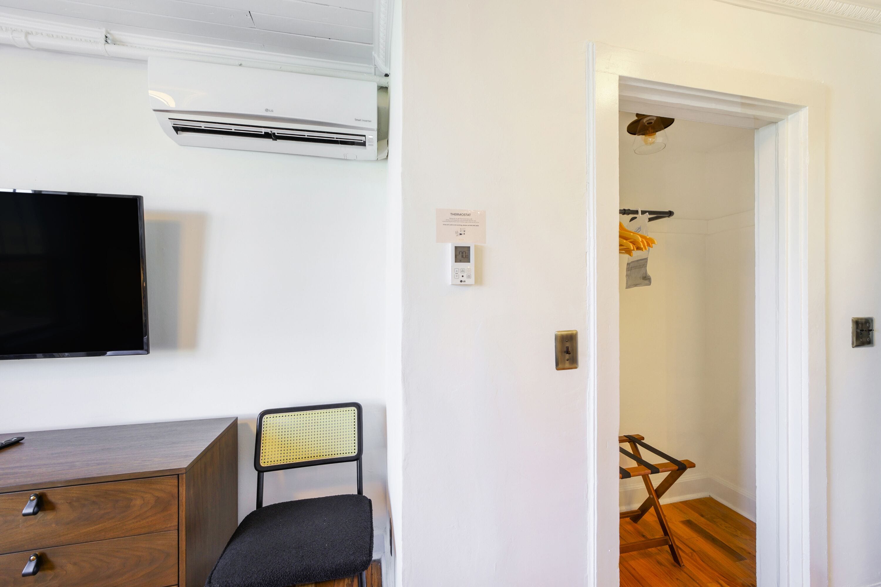 A room with a flat-screen TV, a wooden dresser, a chair, an air conditioning unit, and a door leading to a bathroom with a shower and a towel rack.