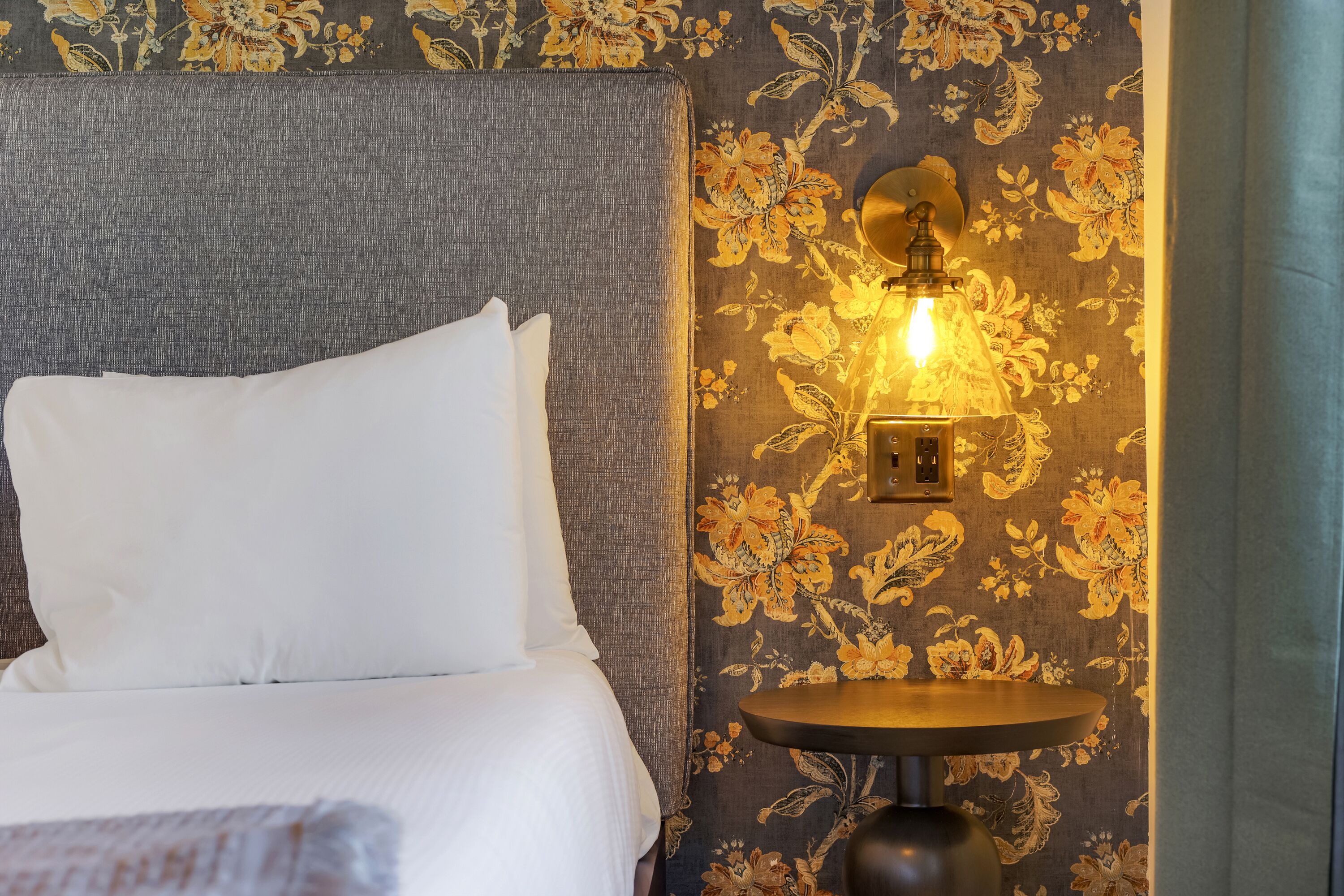 Un lit bien fait, avec du linge de lit blanc, une tête de lit grise et un mur recouvert d’un papier peint à motifs floraux.