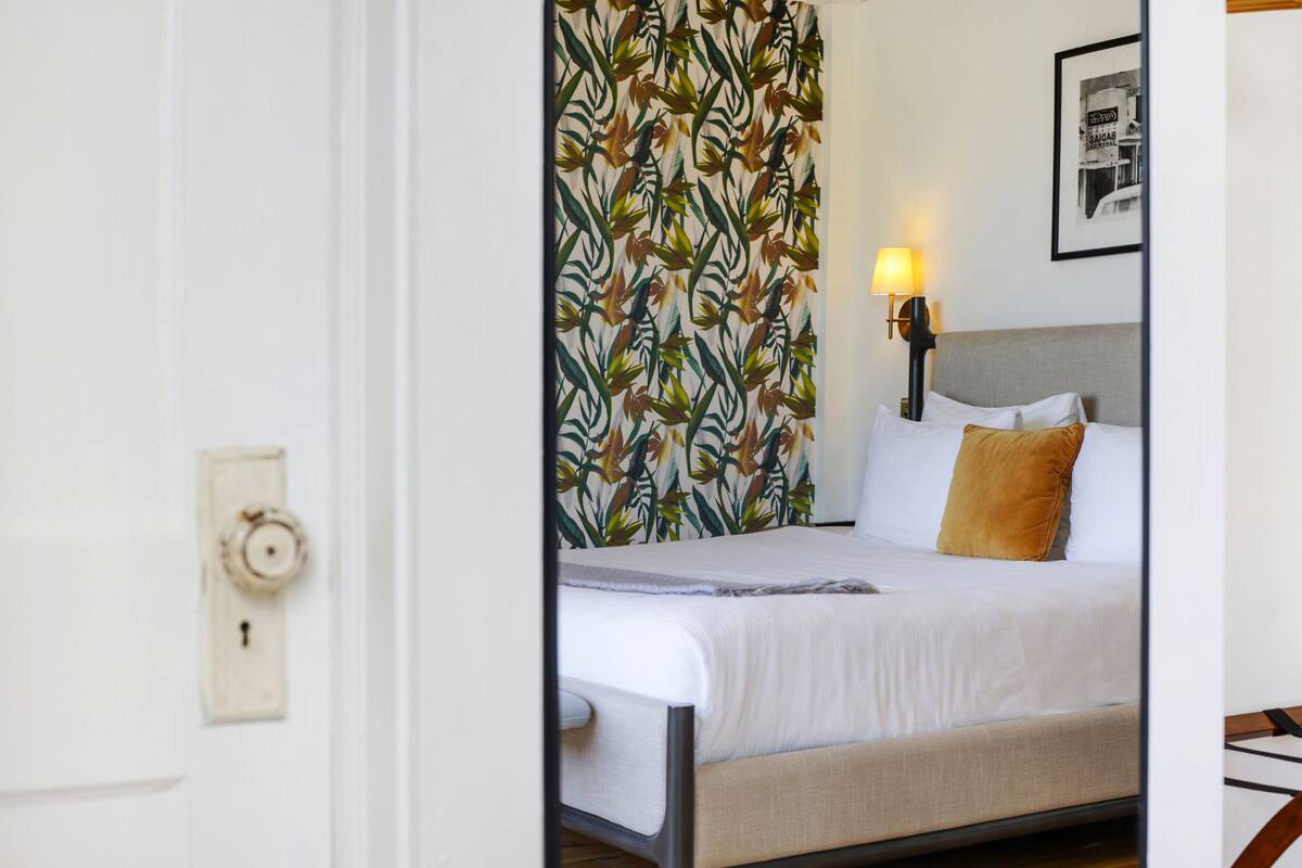 A hotel room with a bed, a nightstand with a lamp, a door with a keyhole, and a patterned curtain.