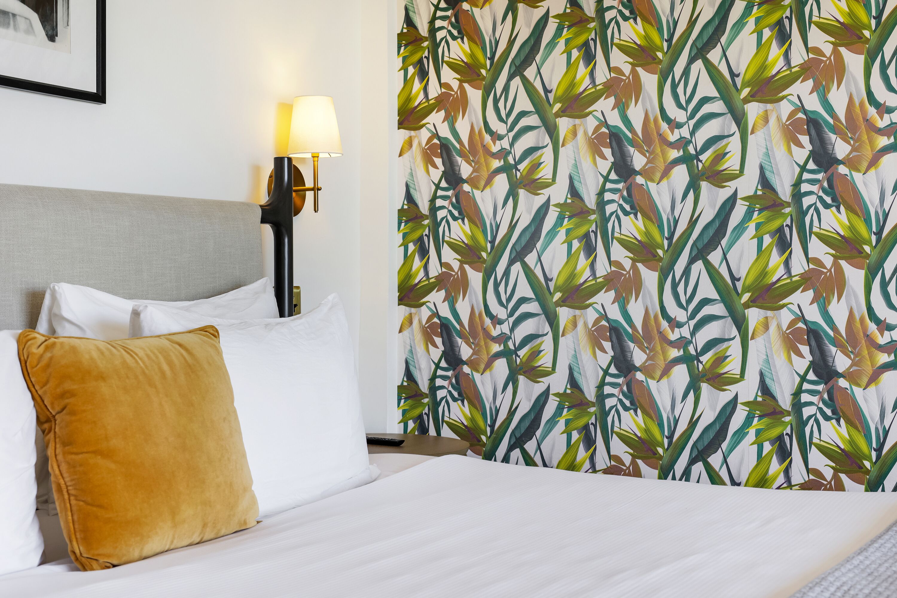 Habitación de hotel con cama, mesita de noche, lámpara y cortina con estampado de hojas tropicales.