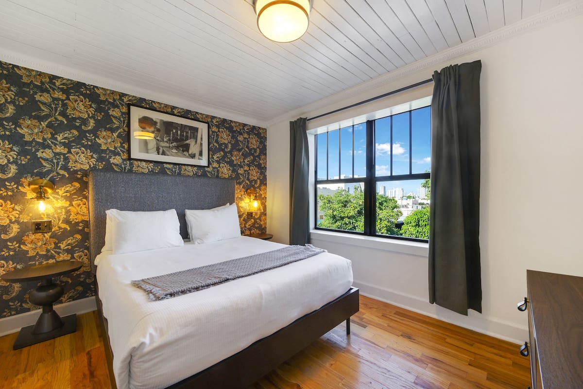 A bedroom with a bed, a nightstand, a wall-mounted lamp, a window with curtains, and a view of greenery outside.