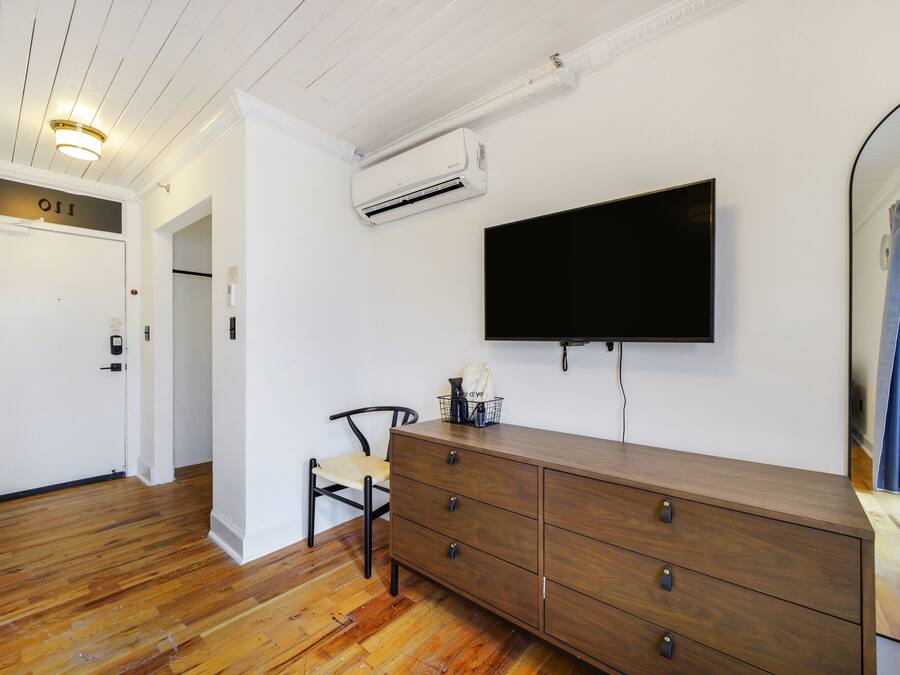 A room with a wooden dresser, a flat-screen TV mounted on the wall, a chair, and an air conditioning unit.