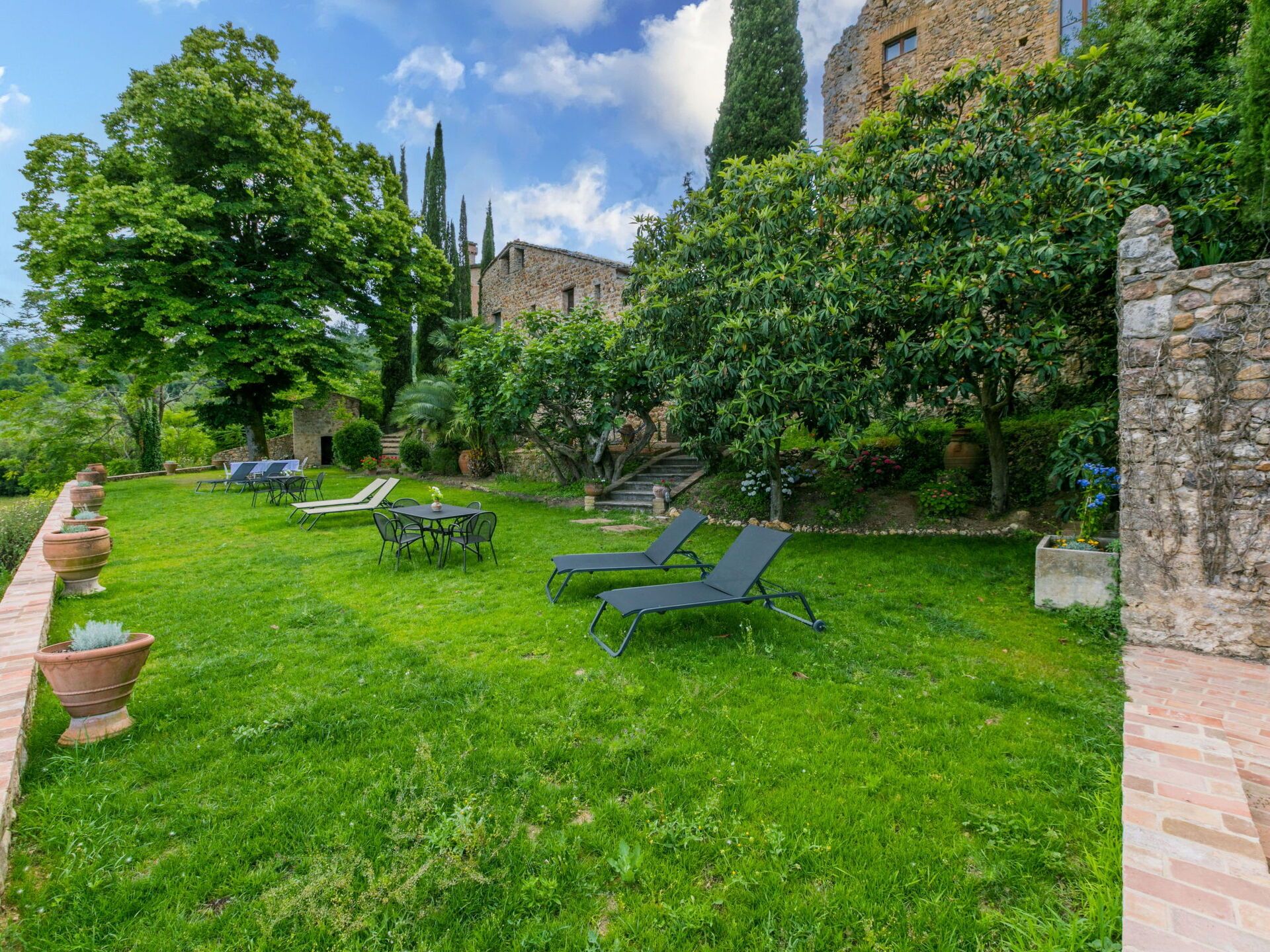 Ein üppiger Garten mit einem Steinhaus, Sitzgelegenheiten im Freien und verschiedenen Bäumen.