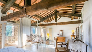 Amplia habitación con techo alto de madera, una cama con colcha blanca, una silla de madera, una mesa con mantel blanco y un aparador con espejo.