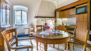 Un comedor con una mesa redonda de madera, seis sillas y una araña. Se ve una cocina con armarios de madera y una estufa al fondo.