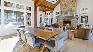 A spacious living area with a large wooden table and comfortable seating, a stone fireplace, and large windows offering a view of the outdoors. - Luxury & Modern Mountain Chalet in The Colony (Park City)
