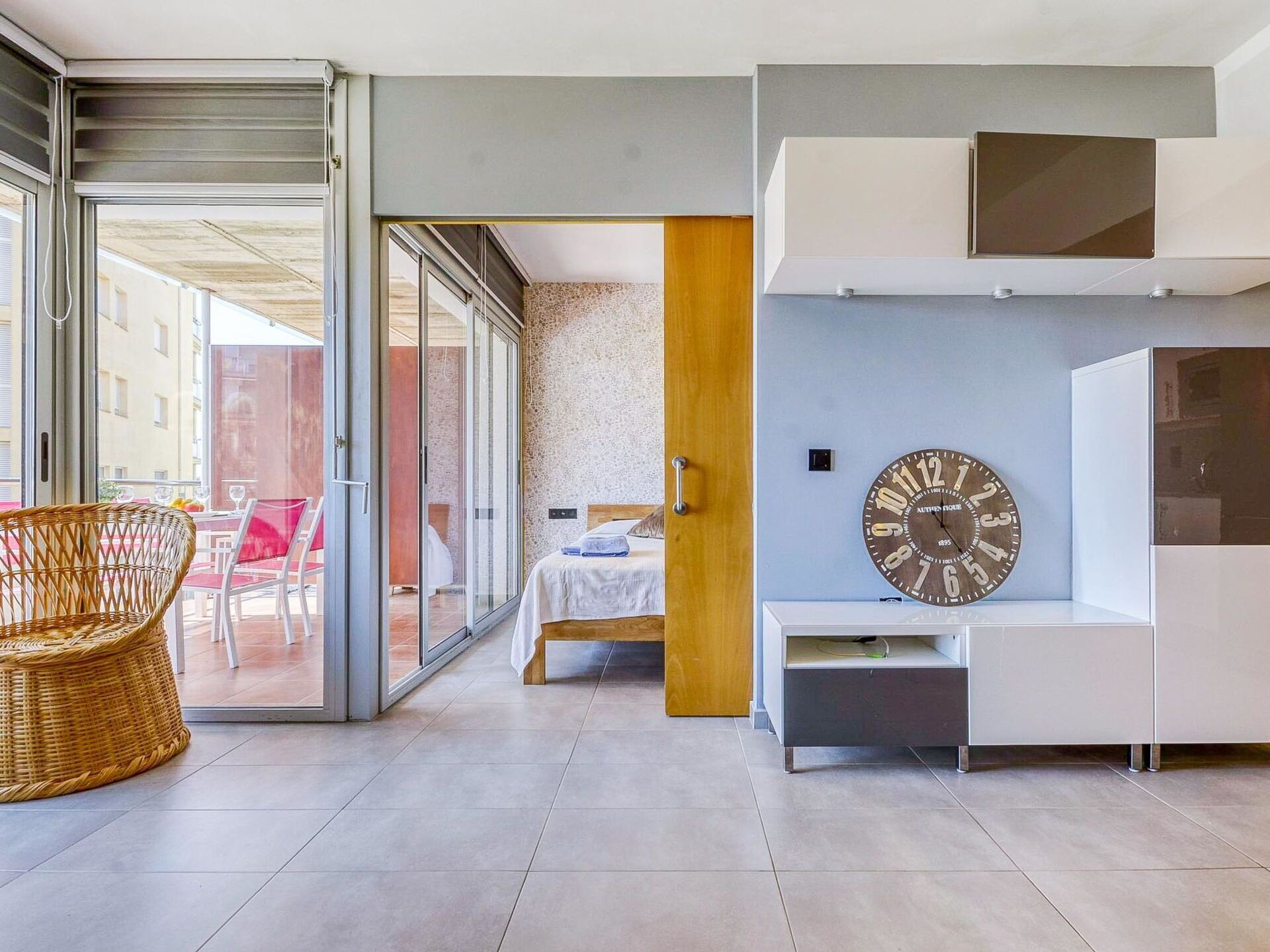 A modern living room with a bed, a large wall clock, and a sliding glass door leading to an outdoor seating area.