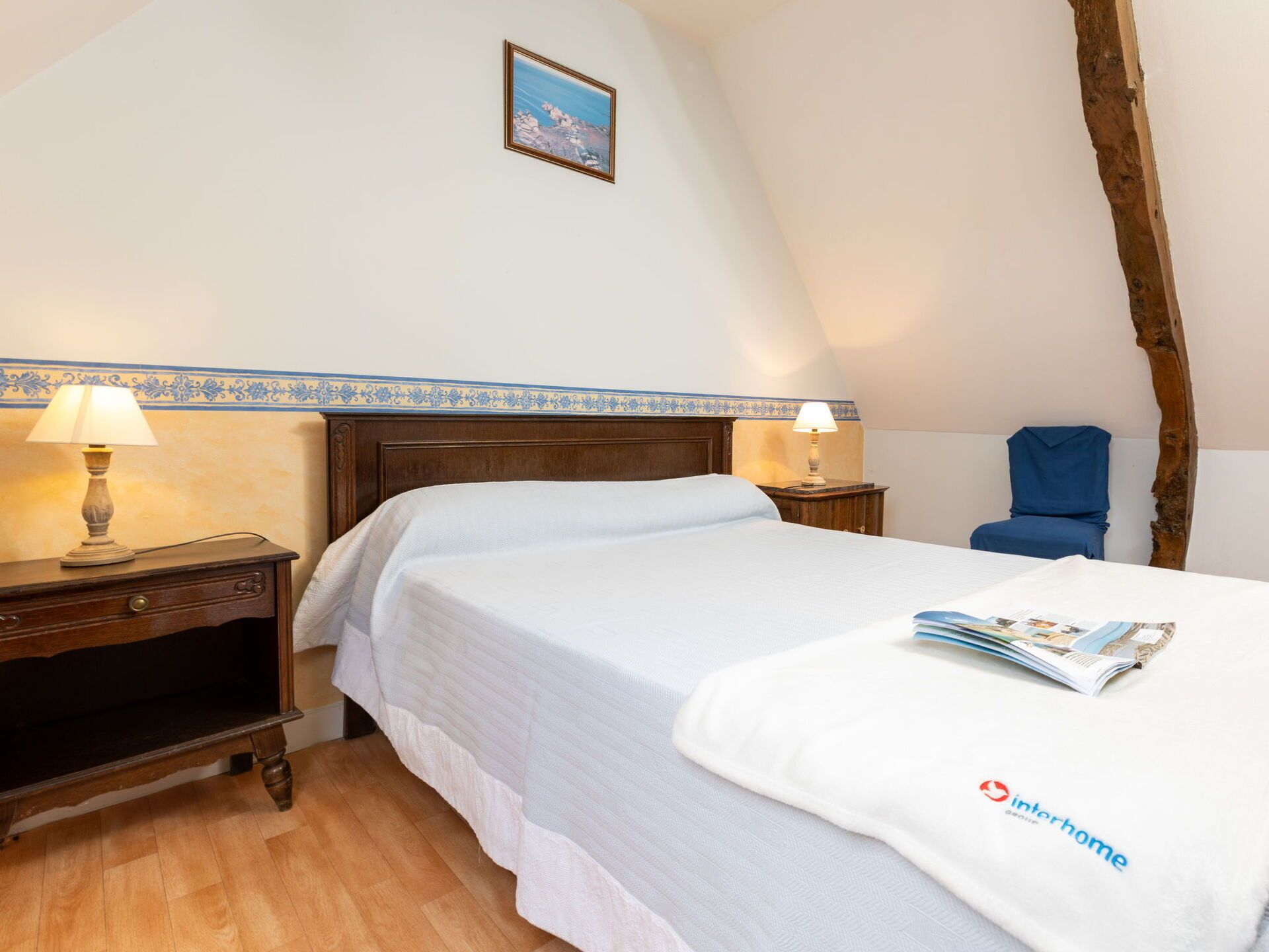 A bedroom with a wooden bed, two bedside tables with lamps, a small chair, a framed picture on the wall, and a magazine on the bed.