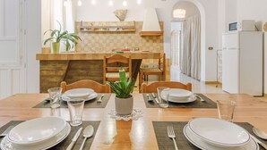 Un coin repas avec une table en bois dressée pour quatre personnes, une cuisine en arrière-plan et un luminaire suspendu.