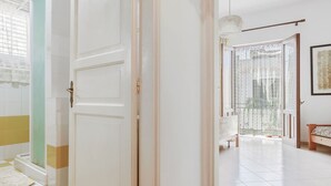 Un couloir avec une porte blanche, un sol à carreaux et une vue sur une pièce où se trouvent un lit et une fenêtre ornée de rideaux à motifs.