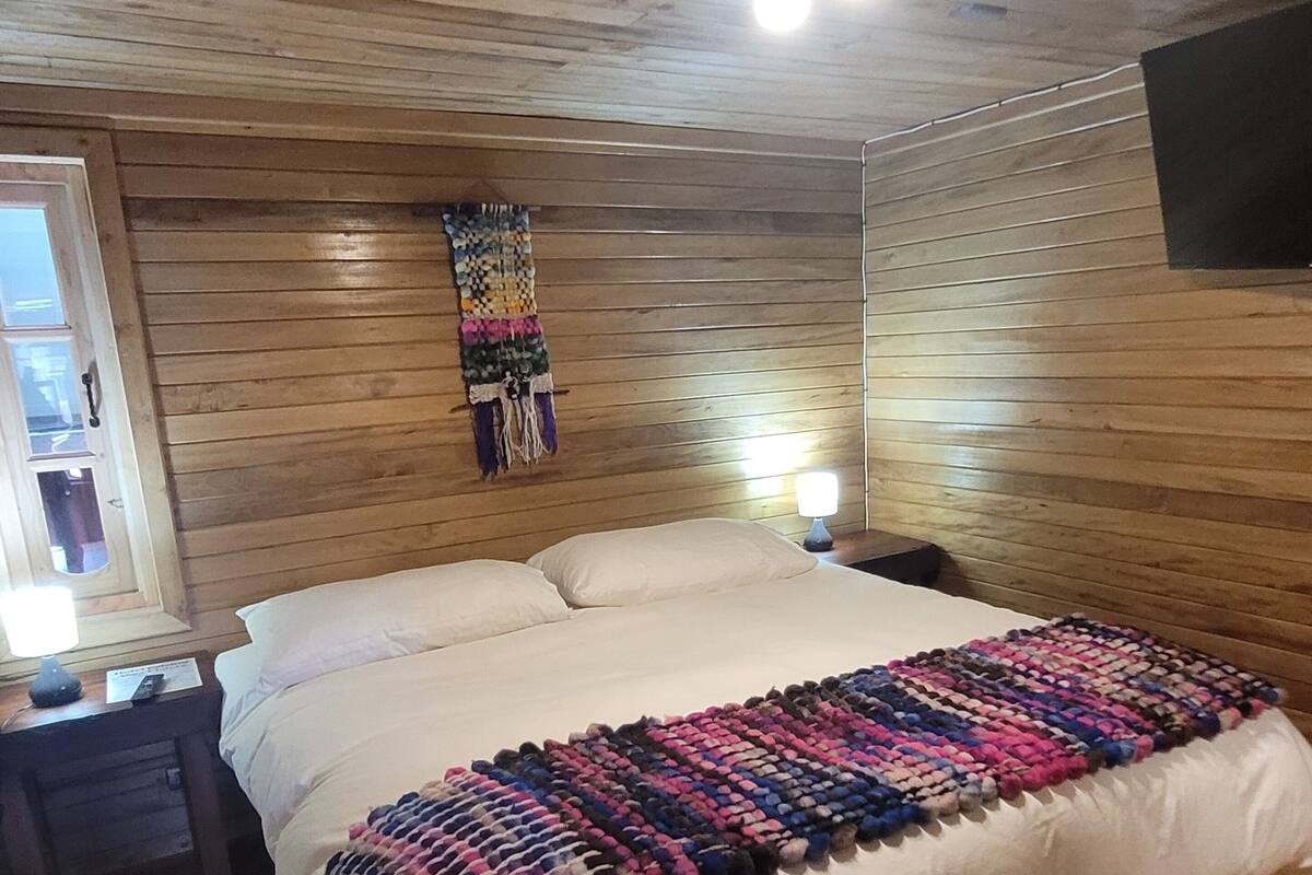 A wooden-paneled bedroom with a large bed, a bedside table, a wall-mounted TV, and a colorful woven blanket.