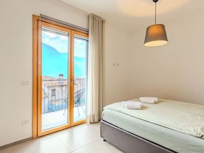 A bedroom with a bed, a hanging lamp, a sliding glass door to a balcony, and a view of buildings and mountains. - Cosy apartment for 5 guests with A/C, WIFI, TV and balcony (Porlezza)