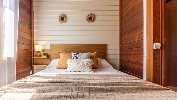 Ein Schlafzimmer mit Holz-Kopfteil, einem Bett mit beiger Bettdecke, zwei Wandleuchten und einem Nachttisch mit Lampe und kleiner Pflanze.