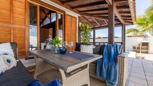Eine überdachte Terrasse mit Korbmöbeln, einem Tisch mit Gläsern und einer Pflanze sowie Blick auf das Meer.